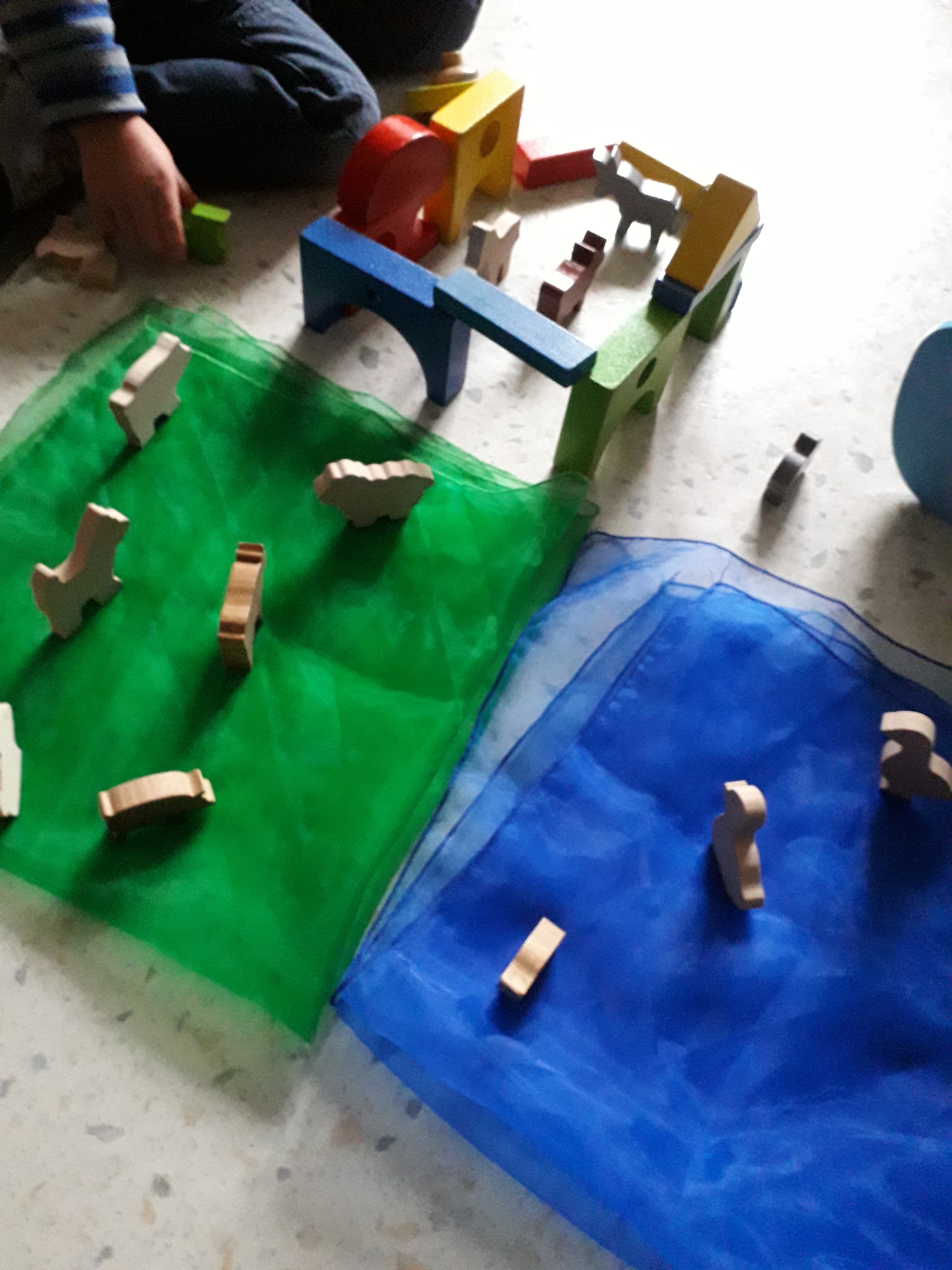 Einige Holz-Bausteine in Haus-Form sind im Viereck aufgestellt, zwischen ihnen stehen kleine Tierchen aus Holz. Daneben liegen ein blaues und ein grünes Tuch, auf denen auch mehrere Tiere stehen. In der oberen linken Ecke sind die Beine und Hände eines Kindes zu sehen, das gerade mit den Holztieren spielt.